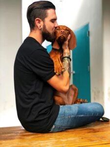 A bearded man sits kissing his dachshund puppy in a casual setting, showing love and companionship.